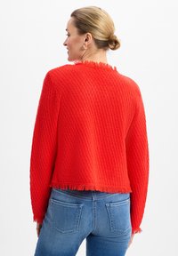 Roter, kürzer geschnittene Pullover mit strukturiertem, gewebtem Muster und fransigem Saum. Lange Ärmel, schlichtes Design und eine lockere Passform. Darunter werden Jeans aus Denim getragen.