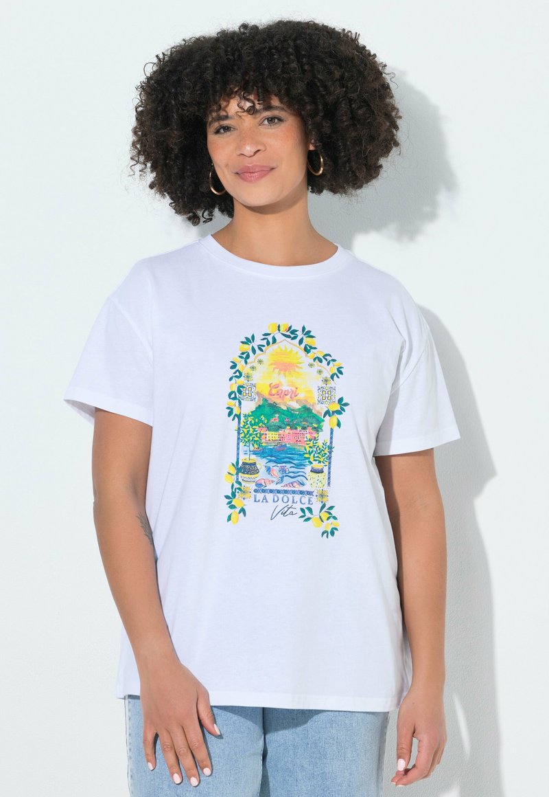 Femme aux cheveux bouclés portant un t-shirt blanc avec une scène côtière italienne colorée et le texte « LADOLCE Vita », associé à un jean bleu clair.