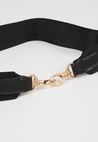 Sangle en tissu noir avec texte en relief, boucle en métal doré et accents en cuir noir sur fond blanc.