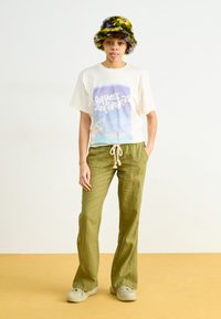 Witte t-shirt met kleurrijke graphic en tekst, gecombineerd met lichtgroene broek met trekkoord. Fluffy emmerhoed in groen, blauw en geel.