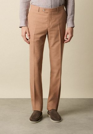 Mężczyzna w beżowych, dopasowanych spodniach, jasnoszarej koszuli zapinanej na guziki i wsuwanych brązowych butach, stojący na szarej podłodze na tle beżowej ściany.