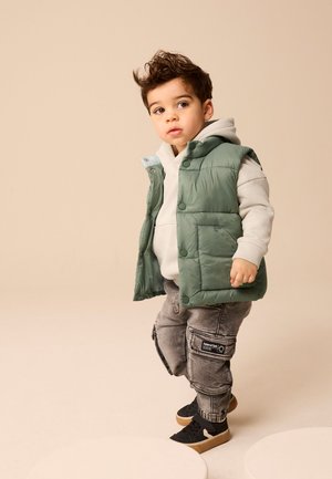 Bambino piccolo con capelli ricci scuri che indossa una felpa con cappuccio grigio chiaro, un gilet imbottito verde, pantaloni cargo grigi e sneakers nere mentre cammina su uno sfondo beige.