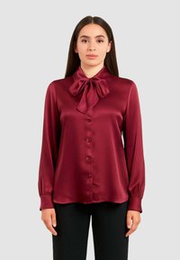 Blusa di raso bordeaux con colletto a fiocco, maniche lunghe e chiusura con bottoni. Presenta una consistenza morbida e un design aderente. Abbinata a pantaloni neri.