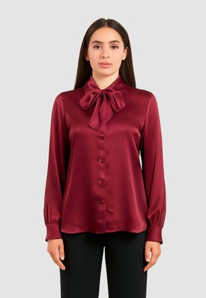 Blusa di raso bordeaux con colletto a fiocco, maniche lunghe e chiusura con bottoni. Presenta una consistenza morbida e un design aderente. Abbinata a pantaloni neri.