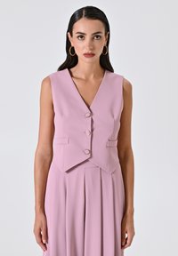 Helles rosa ärmelloses Oberteil mit V-Ausschnitt, drei runden Knöpfen und seitlichen Taschen, kombiniert mit einem plissierten Rock in derselben Farbe.
