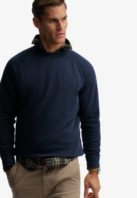 Felpa blu marino con tessuto testurizzato, maniche raglan e piccolo logo. Indossata sopra una camicia a quadri verde, pantaloni beige e un orologio.