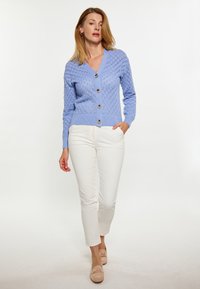Cardigan tricotat de culoare albastru deschis, cu model în diamant, cu decolteu în V și cinci butoane maro. Asortat cu pantaloni albi slim-fit și pantofi slip-on bej.