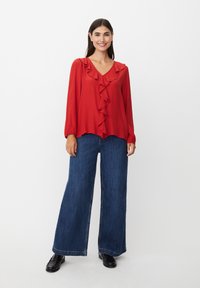Rote Bluse mit Rüschendetails, V-Ausschnitt und langen Ärmeln, kombiniert mit weit geschnittenen Denim-Hosen. Schwarze Schuhe runden das Outfit ab.