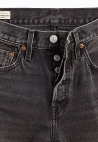 Pantalones vaqueros de mezclilla negra con un diseño clásico de cinco bolsillos, que presentan botones a presión plateados y detalles de costura sutiles, exhibiendo un acabado texturizado.