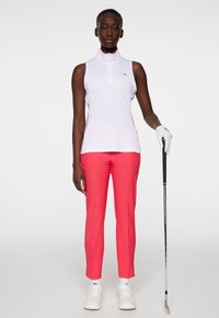 Lättviktig vit ärmlös polo med hög krage och röda accenter, kombinerad med rosa åtsittande byxor. En golfklubba hålls i vänster hand.