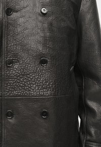 Veste en cuir noir avec une finition texturée, comportant six boutons noirs et un design croisé avec des accents de couture visibles.