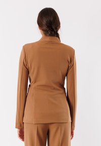 Blazer marrone su misura con un design elegante, caratterizzato da maniche lunghe, colletto alto e una silhouette aderente. Tessuto di consistenza liscia.