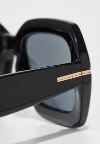 Crne sunčane naočale s pravokutnim okvirima, sivo zatamnjene leće i zlatni detalji na sljepoočnicama. Gladak plastični materijal s sjajnim finishem.