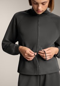 Jachetă zip-up de culoare gri închis, cu textură ribbed și guler înalt. Are șnururi reglabile la talie și mâneci cu o croială lejeră.