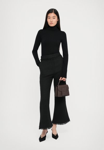 Black turtleneck top paired with high-waisted black pants featuring fringe accents. Model holds a small dark brown handbag. Black heeled shoes.