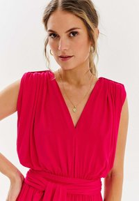 Różowa sukienka bez rękawów z dekoltem w kształcie litery V, plisowanym biustem i ściągniętym w talii. Zawiera zawiązywany pasek oraz naszyjnik z małym wisiorkiem.