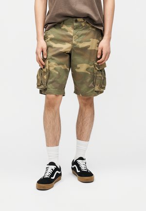 Person trägt grüne und braune Camouflage-Cargoshorts, weiße Socken und schwarze Sneakers mit weißen Schnürsenkeln und steht vor einem einfarbigen Hintergrund.