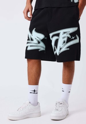 Man draagt zwarte korte broek met grote witte graffiti-print, witte sokken met zwarte logo's en witte Nike-sneakers, staand tegen een effen achtergrond.
