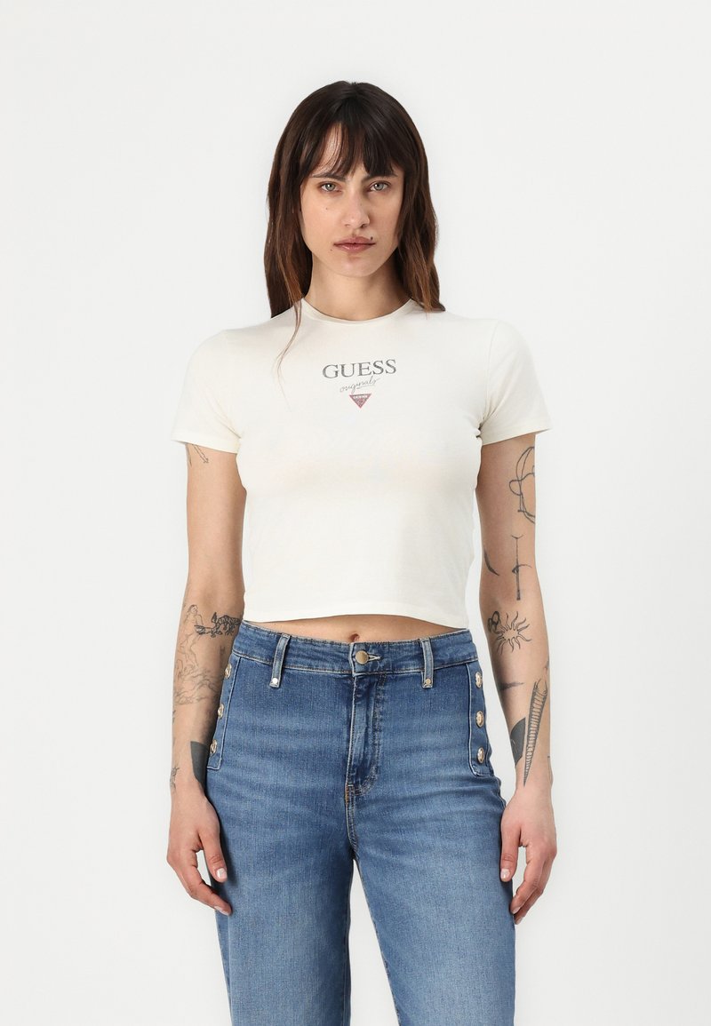 Kermansävyinen lyhythihainen crop-top, jossa on "GUESS"-logo ja kolmiomainen muotoilu, yhdistettynä korkeavyötäröisiin sinisiin farkkuihin, joissa on nappikoristeet.