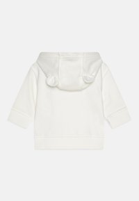 Balts kapuci sūdzis ar apaļiem ausu līdzīgiem akcentiem uz kapuces, garām piedurknēm un ribinātu apakšmalu. Izgatavots no mīksta, teksturēta auduma.