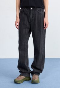 Jeans a gamba larga a strisce nere realizzati in denim, con tasche frontali, patta con bottone e una vestibilità comoda, abbinati a sneakers marroni e verdi.