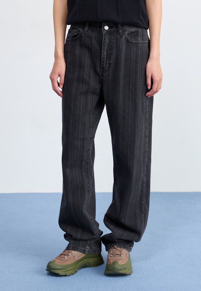 Jeans a gamba larga a strisce nere realizzati in denim, con tasche frontali, patta con bottone e una vestibilità comoda, abbinati a sneakers marroni e verdi.