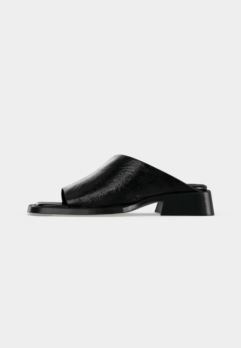 Μαύρο δερμάτινο σανδάλι slides με ανοιχτά δάχτυλα, χαμηλό τακούνι και λείο υφή επάνω μέρος, απεικονίζεται σε απλό ανοιχτό γκρι φόντο.