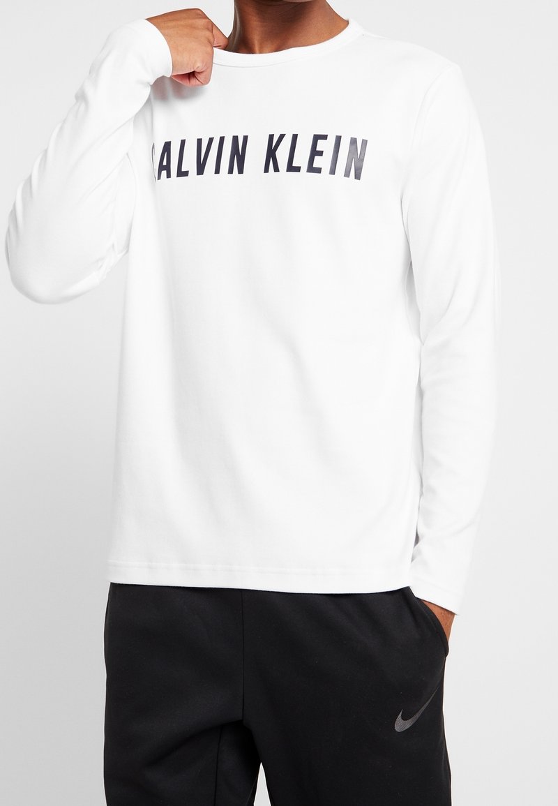 Osoba v bílé košili s dlouhými rukávy s textem "CALVIN KLEIN" a černých kalhotách Nike, s pěsťí zvednutou u ramene a rukou v kapse.