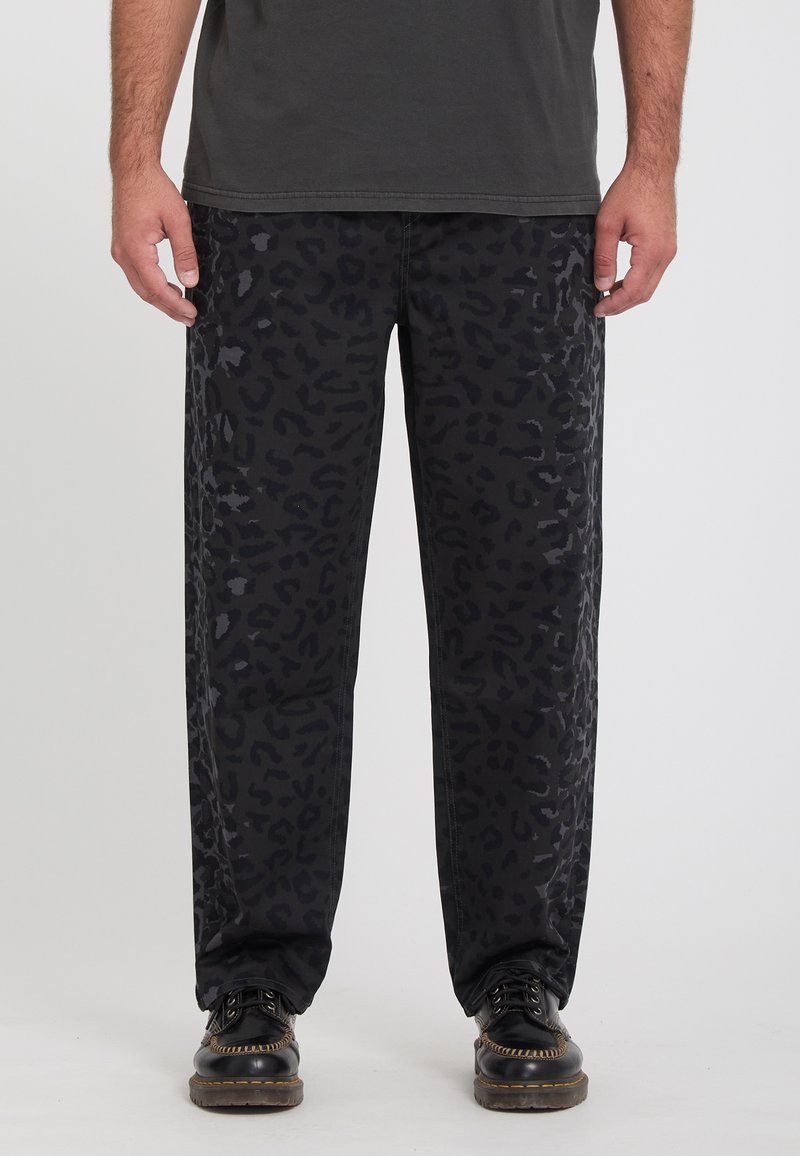 Homme portant un pantalon noir avec un imprimé léopard gris foncé et des chaussures en cuir noires, debout devant un fond uni.