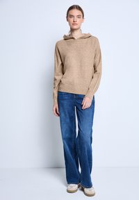 Beige gestrickter Hoodie mit lässigem Schnitt und Kordelzug, kombiniert mit blauen Jeans im geraden Schnitt und beigen Sneakers, vor einem schlichten Hintergrund getragen.