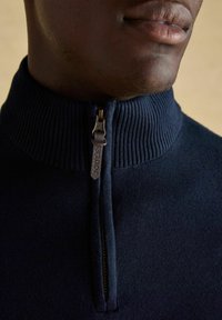 Zbliżenie osoby w granatowym swetrze zapinanym na zamek błyskawiczny z prążkowanym kołnierzem oraz z suwakiem oznaczonym napisem "JOULES."