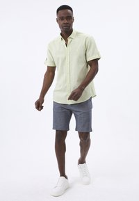 Hellgrünes Kurzarmhemd mit Button-Down-Kragen und aufgerollten Ärmeln, kombiniert mit dunkelgrauen Shorts und weißen Sneakers.