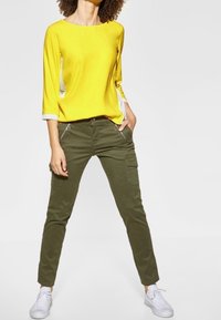 Gul tröja med trekvartslånga ärmar och vita detaljer, i kombination med olivgröna cargobyxor med sidfickor, bärs med vita sneakers.