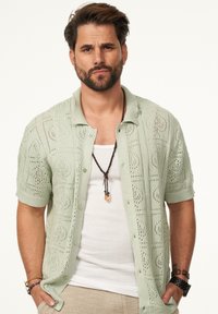 Hellgrünes, gestricktes Kurzarmhemd mit Knopfleiste und Durchbrochenmuster, ausgestattet mit einem Kragen und strukturiertem Stoff. Über einem weißen Tanktop getragen.