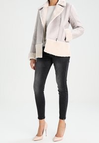 Chaqueta de pelo sintético gris claro con textura acolchada, ribete crema y cremallera lateral. Combinada con jeans ajustados negros y tacones altos rosa claro.