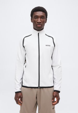 Mann trägt eine weiße Jacke mit Reißverschluss und schwarzem Besatz sowie dem "BOSS"-Logo, kombiniert mit hellbraunen Hosen.