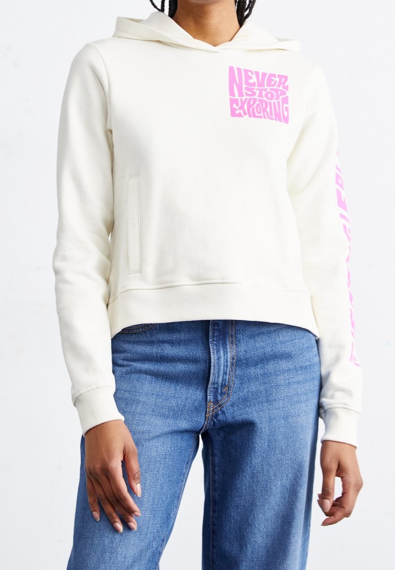 Personne portant un sweat à capuche blanc avec le texte rose « Never Stop Exploring » sur la poitrine et la manche, associé à un jean bleu.