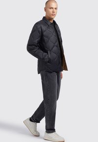 Sort polstret jakke med et quiltet mønster, trykknaplukning og sidelommer; båret over en sort skjorte og parret med grå jeans og hvide sneakers.