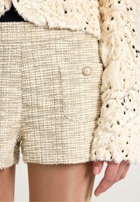 Beige tweed shorts med en struktureret overflade og en lille sidelomme. Pyntet med en perleknap og parret med en fluffy gylden jakke med paljetter.