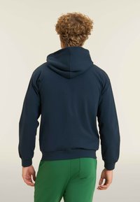 Uomo con capelli ricci che indossa un hoodie blu navy e pantaloni verdi, in piedi rivolto di spalle contro uno sfondo chiaro e uniforme.