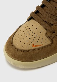 Rjavi semofor s perforiranim prstom, oranžna Nike značka in bež gumijast podplat. Ima rjave vezalke in tekstilne detajle.