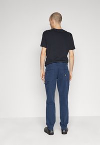 Pantaloni cargo in cotone blu navy con tasche laterali e toppe posteriori, abbinati a una t-shirt nera a girocollo e scarpe nere, visti da dietro.