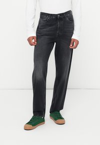 Dunkelgraue Jeans mit geradem Bein, sichtbaren Nähten, fünf Taschen und einem Knopfverschluss, kombiniert mit grünen Sneakers.