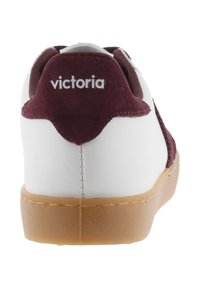 Vit sneaker med en vinröd mockahälkappa. Har en gummisula och varumärket "victoria" tryckt på baksidan.