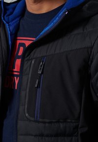 Musta takki, jossa sininen fleece-vuori, vetoketjuyksityiskohdat ja teksturoitu kangas. Sisältää rintataskun, jossa on vetoketju ja joka on erivärinen.