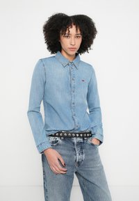 Svetlomodrá denimová košeľa s gombíkmi, golierom a červeným logom. Nosí sa s vybledlými modrými denimovými džínsami a čiernym opaskom s cvočkami.