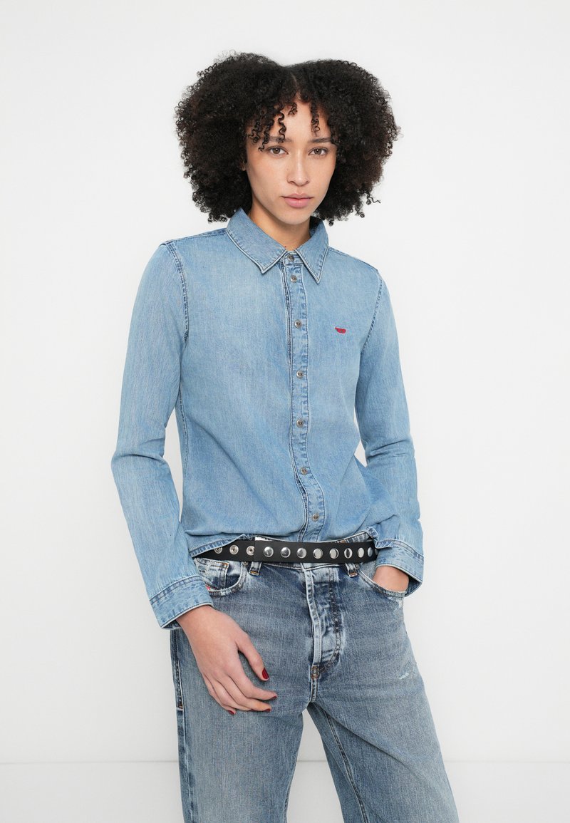 Lyseblå denimskjorte med knapper, krave og rødt logo-detalje. Bæres med falmede blå denimjeans og et sort bælte med nitter.