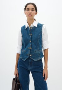 Denim vest met een slim fit ontwerp, blauwe kleur, vijf metalen knopen en twee voorzakken. Draagt over een wit overhemd met kraag.