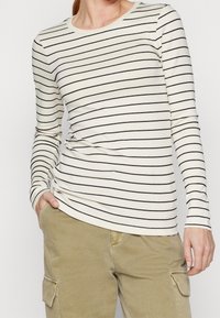 Person som bär en krämfärgad långärmad skjorta med tunna svarta horisontella ränder och khaki cargo-byxor, med handen i fickan, beskuren vid nacken.