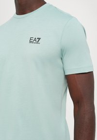 Mintgrønn kortermet t-skjorte laget av myk stoff, med en svart EA7 Emporio Armani-logo på brystet. Jevn tekstur og kroppsformet design.